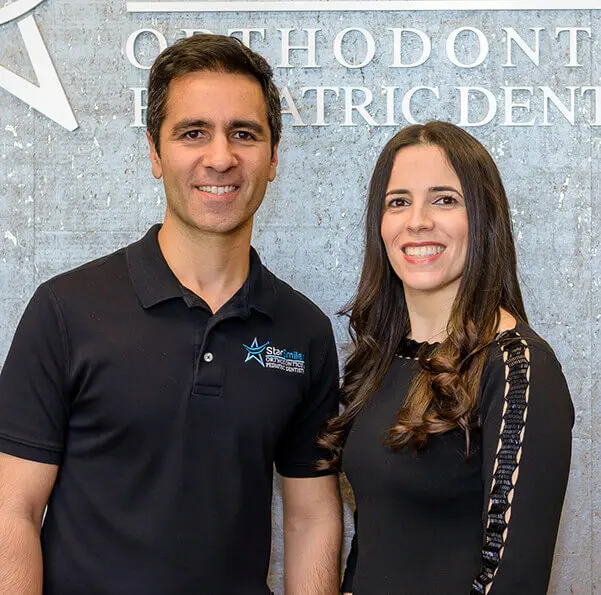 At Star Smile Orthodontics in Bloomingdale IL, Drs. Andre Jham and Raquel Martuchi Jham—a male and female dentist—stand together smiling in front of an orthodontic and pediatric dentistry sign.