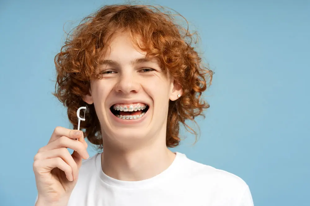 A teen with curly red hair and braces smiles while holding a dental floss pick against a plain blue background, showcasing Star Smile Orthodontics in Bloomingdale, IL.