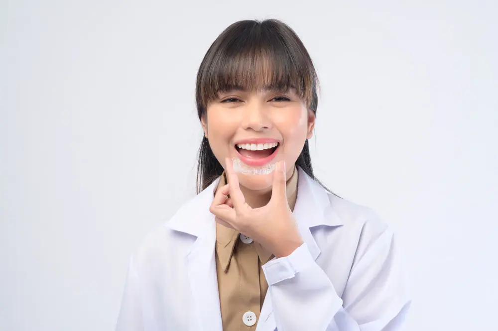 At Star Smile Orthodontics in Bloomingdale IL, a person in a white coat smiles while holding Invisalign® Clear Aligners in front of their teeth against a plain background.