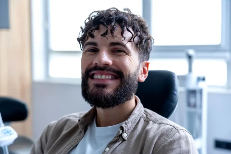 Smiling confidently in a brightly lit office, a man with curly hair and a beard sits in the dental chair at Star Smile Orthodontics in Bloomingdale IL, proud of his choice to get clear braces.