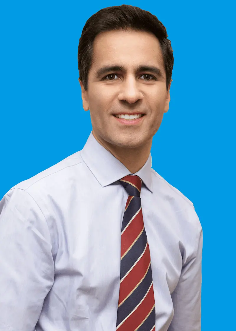 Dr. Andre C. Jham, a board-certified orthodontist at Star Smile Orthodontics in Bloomingdale IL, confidently smiles in a light blue dress shirt and red and blue striped tie against a solid blue background.