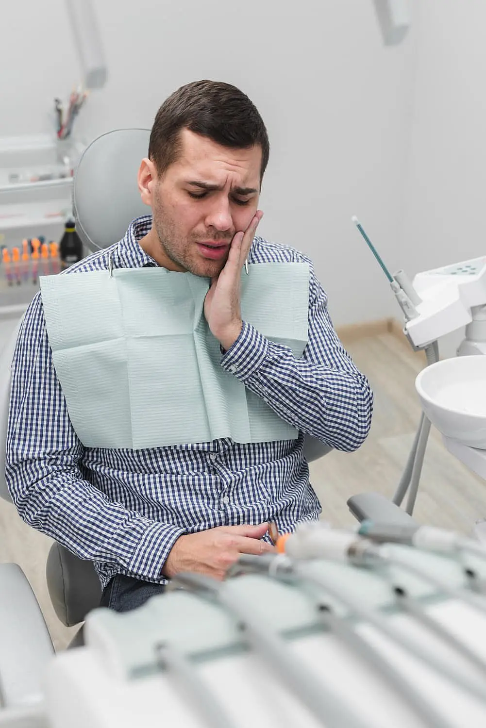 A man sitting in a dental chair, wearing a checkered shirt and dental bib, holds his cheek in discomfort, possibly indicating tooth pain. Dental equipment surrounds him, hinting at the possibility of upcoming jaw surgery in Bloomingdale and Naperville, IL.