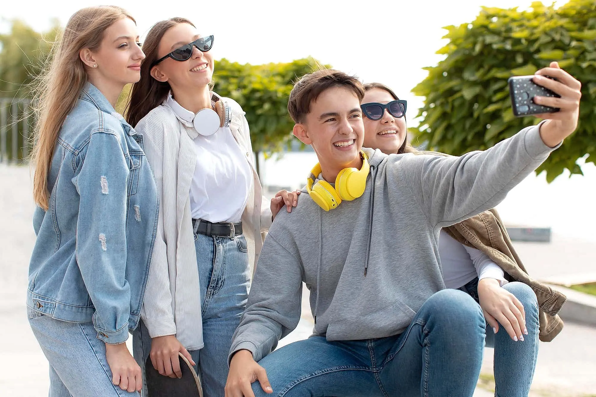 A group of four young people, outdoors in Bloomingdale and Naperville, IL. Chat about different types of braces while taking a selfie—one holds the phone, two wear headphones around their necks, and trees with a walkway are visible behind them. The scene is linked to Star Smiles Orthodontics and Pediatric Dentistry.