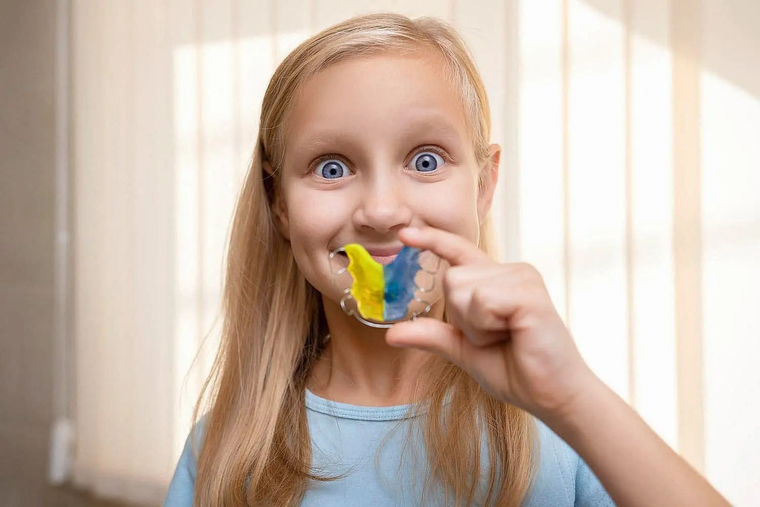 A young girl with long blonde hair proudly smiles indoors, showing off a colorful yellow-and-blue striped retainer—thanks to the expert care at Star Smiles Orthodontics and Pediatric Dentistry in Bloomingdale and Naperville, IL.