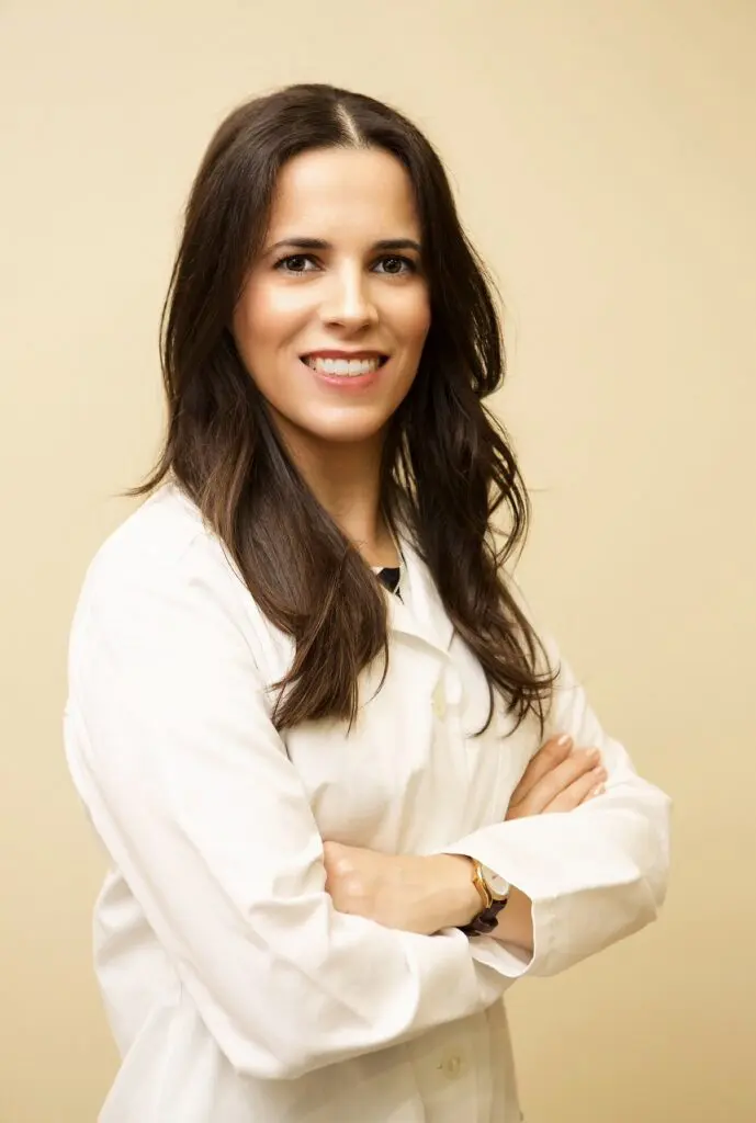 At Star Smiles Pediatric Dentistry in Bloomingdale or Naperville, IL, Dr. Raquel M. Jham, DDS, is pictured with long brown hair and a warm smile, confidently wearing a white lab coat with her arms crossed against a beige background.