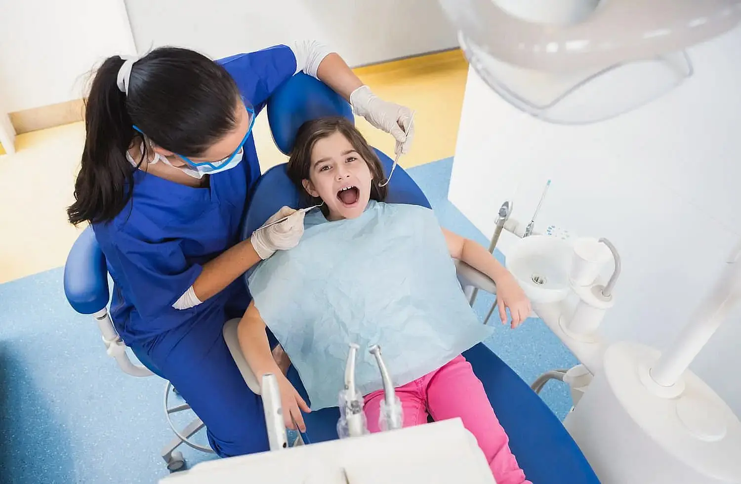 A children's orthodontist examines a child in a dental chair. The young patient wears a bib and has her mouth open, ready for the check-up. The orthodontist is dressed in blue scrubs and gloves, skillfully using dental tools to ensure everything is in order in Bloomingdale and Naperville, IL.