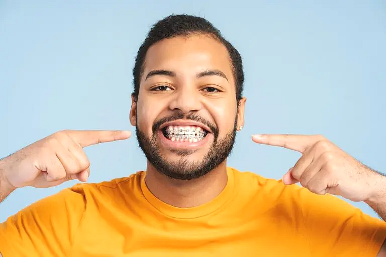 A man in a yellow shirt smiles and points at his adult metal braces against a plain blue background, representing Star Smiles Orthodontics and Pediatric Dentistry in Bloomingdale and Naperville, IL