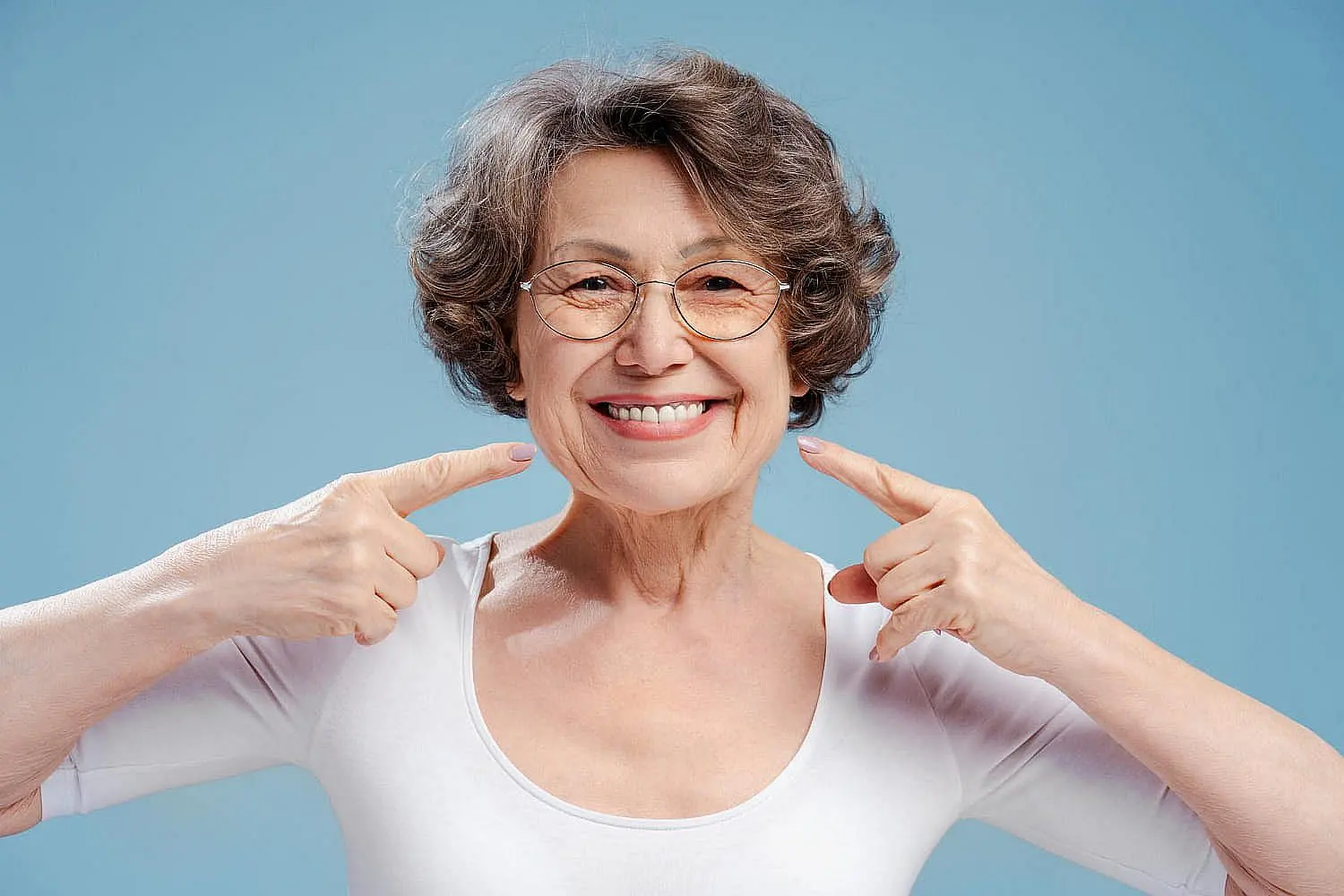 A smiling older woman with short gray hair and glasses points to her teeth against a plain light blue background, highlighting the benefits of orthodontic insurance plans at Star Smiles Orthodontics and Pediatric Dentistry in Bloomingdale and Naperville, IL.