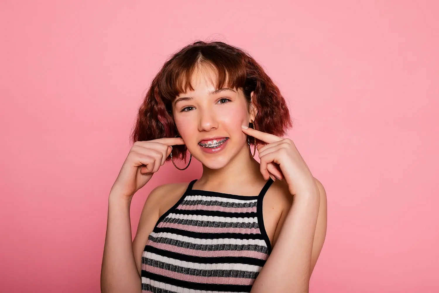 A teen girl with short brown hair and braces stands in front of a solid pink background, wearing a striped sleeveless top. She smiles and points to her cheeks, highlighting her Invisalign vs Braces experience at Star Smiles Orthodontics and Pediatric Dentistry in Bloomingdale and Naperville, IL.