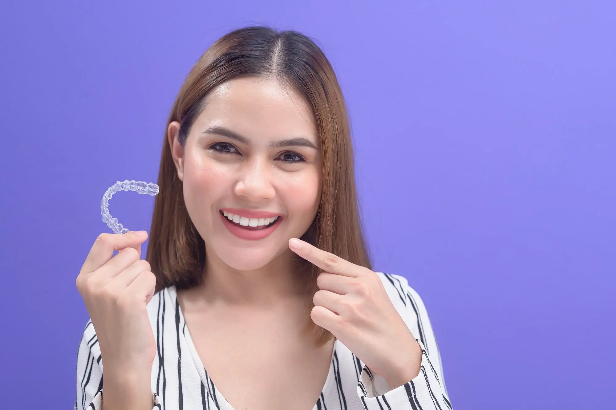 A smiling woman proudly presents a clear dental aligner Invisalign between their fingers, reminiscent of modern braces, in a bright room in Bloomingdale, IL.