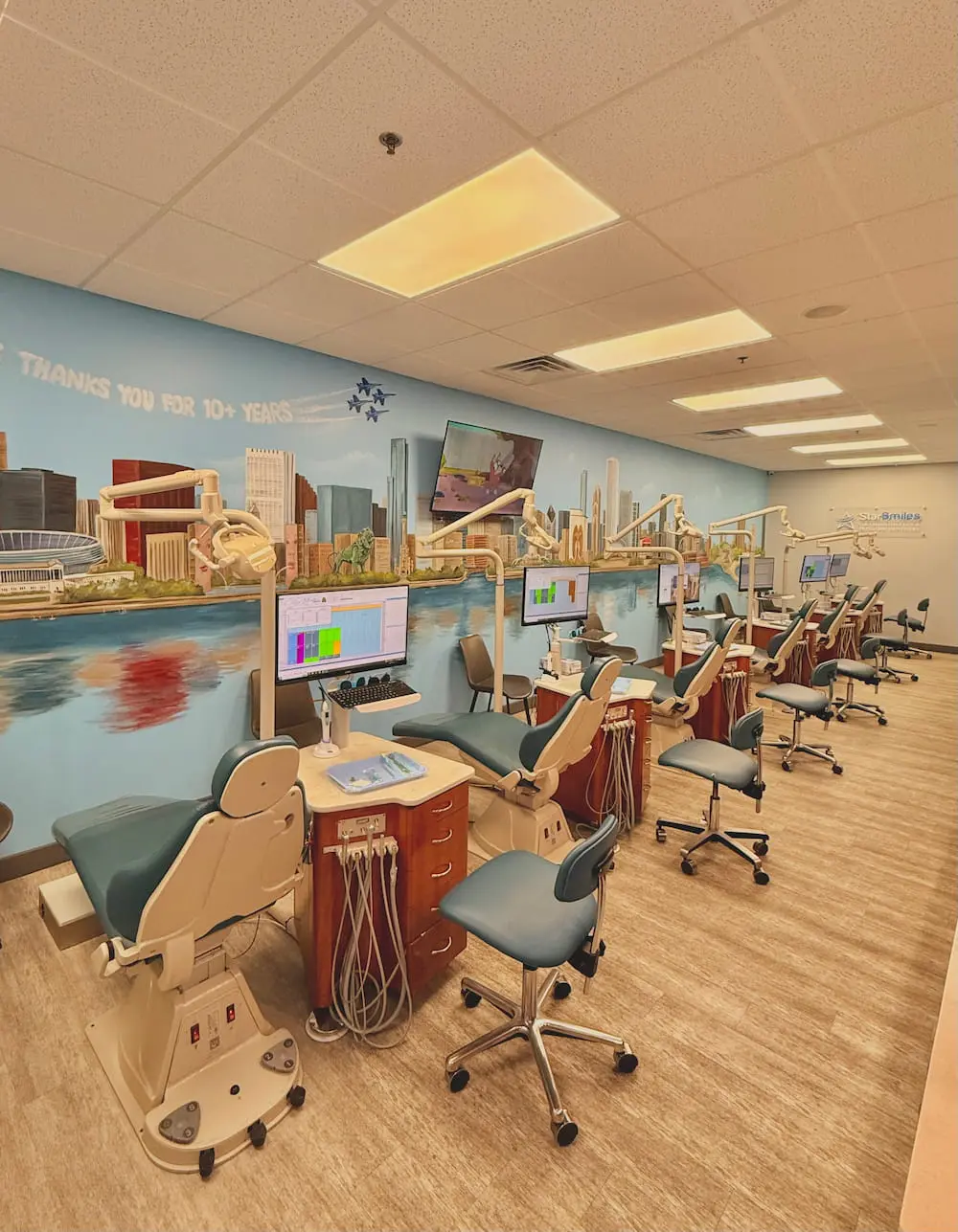 At Star Smiles Pediatric Dentistry in Bloomingdale, IL, a row of dental chairs and workstations faces computer monitors beneath a vibrant cityscape mural in a brightly lit clinic.
