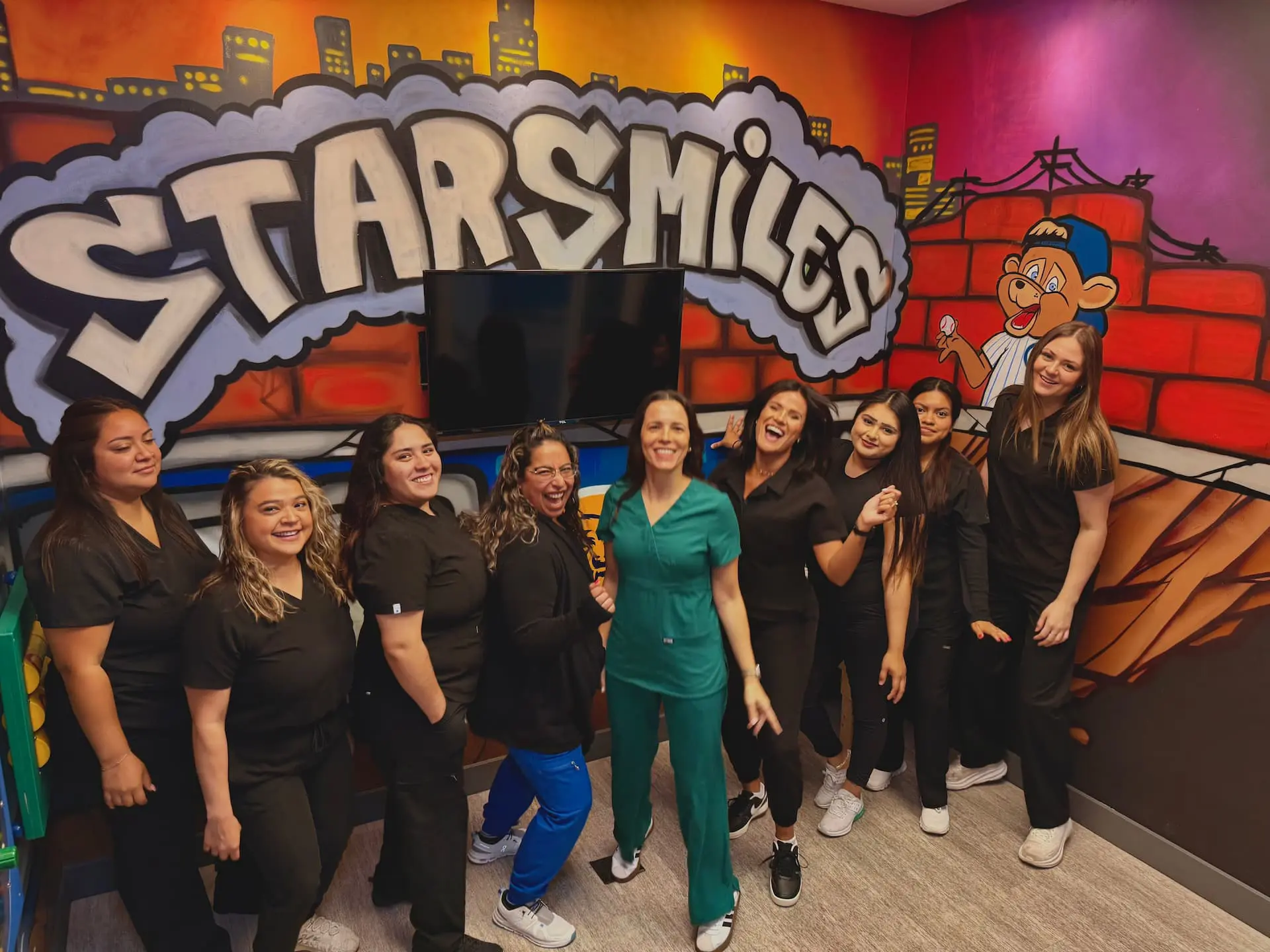 Inside a dental office in Bloomingdale or Naperville, IL, ten women—mostly dressed in black scrubs—smile and pose in front of a colorful mural featuring "Star Smiles Pediatric Dentistry.